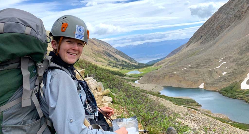a person wearing a helmet and carrying a backpack smiles for a photo in front of an alpine lake